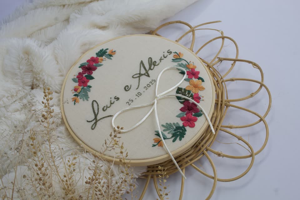 Porta-aliança com os nomes do casal em letra cursiva e flores coloridas bordadas, com laço para as alianças