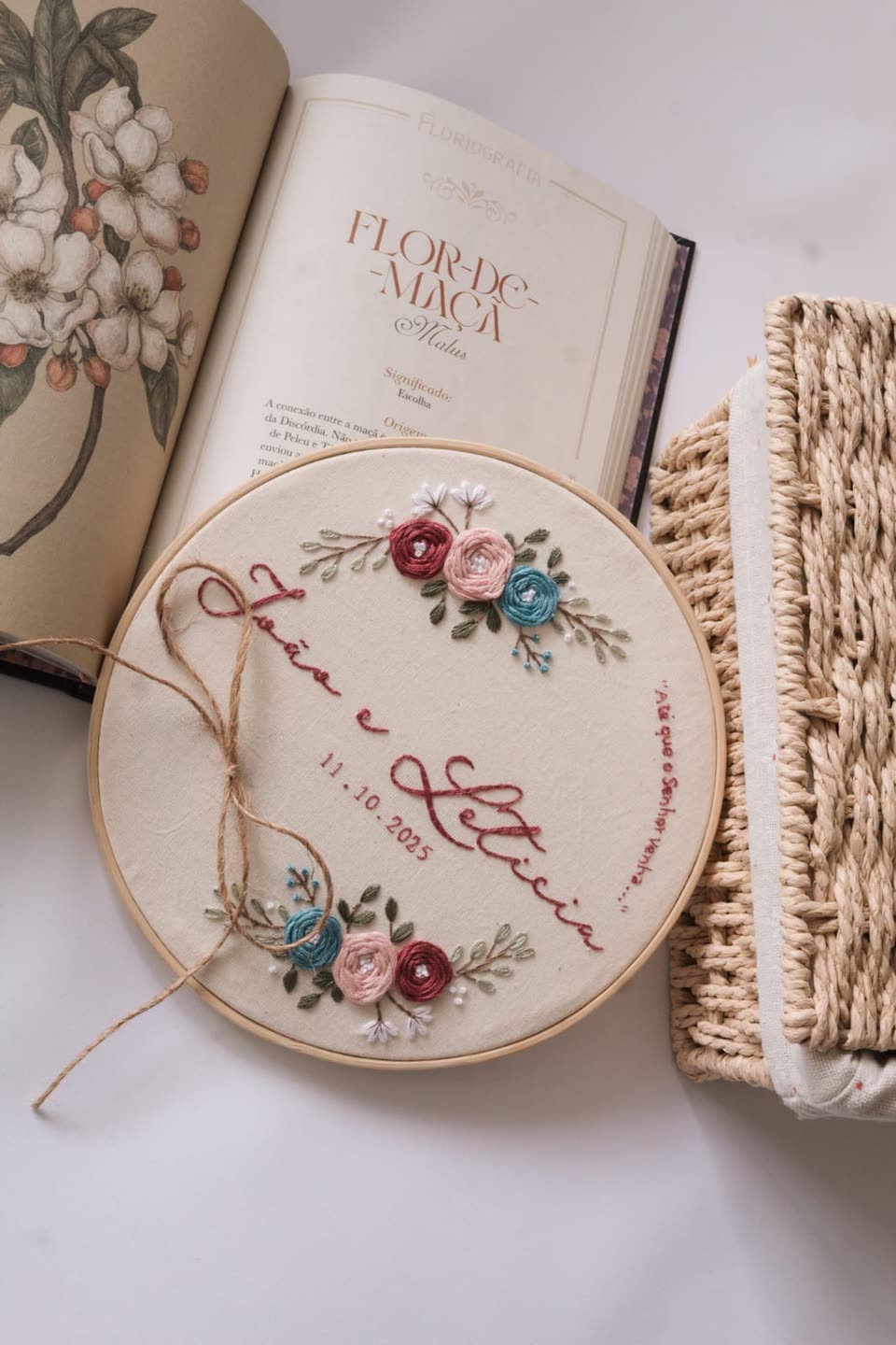 Porta-aliança com flores rosa e azuis bordadas e os nomes do casal em letra cursiva, ao lado de um livro aberto
