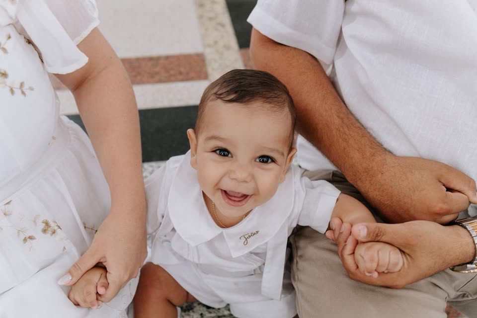 Bebê sorrindo de camisa branca com golinha bordada, no colo dos pais